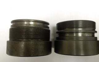 Two black metal fittings on a white surface in a machine shop.